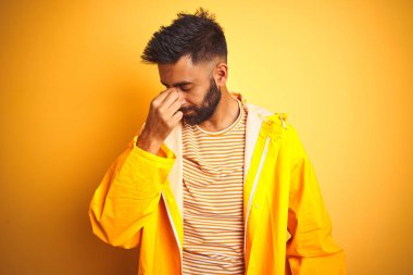 İzole edilmiş sarı arka planda duran yağmurluk giyen genç Hintli adam yorgun burnu ve gözleri ovuşturarak yorgunluk ve baş ağrısı hissediyor. Stres ve hüsran kavramı.