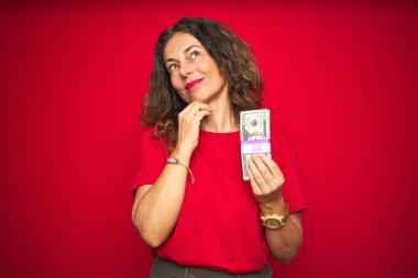 Orta yaşlı bir kadın kırmızı izole edilmiş arka planda bir sürü dolar tutuyor. Ciddi bir yüz ifadesi var. Soru sormayı düşünüyor, kafası karışık bir fikir.