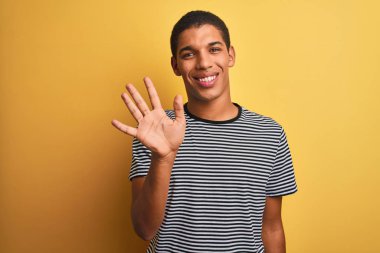 Sarı arka planda donanma çizgili tişört giyen genç yakışıklı Arap adam beş numarayı gösterirken kendinden emin ve mutlu bir şekilde gülümsüyor..