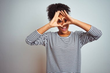 Siyah çizgili tişört giyen Afro-Amerikalı bir kadın izole edilmiş beyaz arka planda durup el ve parmaklarıyla kalp şeklinde işaret diliyle gülümsüyor.