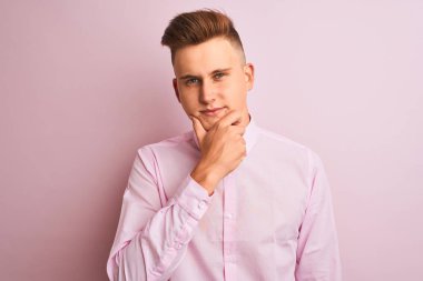 Soyutlanmış pembe arka plan üzerinde duran zarif gömlekli genç yakışıklı işadamı kameraya kollarını kavuşturup gülümseyerek ve çenesini kaldırarak bakıyor. Olumlu düşünüyorum..