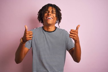 Rasta desenli tişörtlü Afro adam izole edilmiş pembe arka plan başarısı tabelasının üzerinde duruyor. Elleriyle olumlu jest yapıyor, gülümsüyor ve mutlu. Neşeli ifade ve kazanan jest.