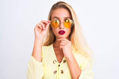 Sarı t-shirt ve güneş gözlüğü takan genç ve güzel bir kadın izole edilmiş beyaz arka plan üzerinde ciddi bir yüz soru hakkında düşünüyor, çok karışık bir fikir.