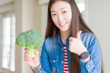 Yeşil brokoli yiyen güzel bir Asyalı kadın gülümsüyor. Tamam işareti yapıyor, parmaklarıyla başparmağını kaldırıyor, mükemmel bir işaret.