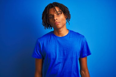 Rastalı tişörtlü Afro-Amerikalı bir adam izole edilmiş mavi arka planda dikiliyor yüzünde ciddi bir ifadeyle rahatlıyor. Basit ve doğal bir şekilde kameraya bakıyorsun..