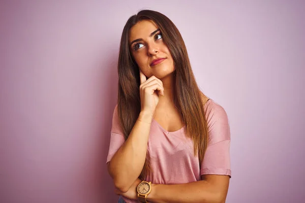 若い美しい女性のTシャツの上に立って孤立したピンクの背景の上に立って質問については 集中的な表現を考えて顎を手にした 思慮深い顔で微笑んだ 疑いの概念 — ストック写真