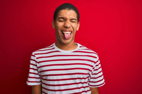 年轻英俊的阿拉伯男子穿着条纹T恤 披着孤立的红色背景 带着滑稽的表情伸出舌头 情感概念 — 图库照片