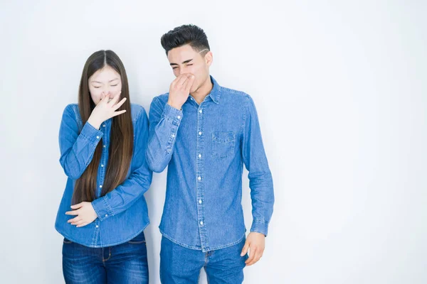 Beautiful young asian couple over white isolated background smelling something stinky and disgusting, intolerable smell, holding breath with fingers on nose. Bad smells concept.