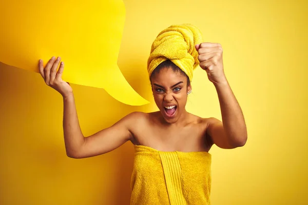 Afro woman wearing towel after shower holding speech bubble over isolated yellow background annoyed and frustrated shouting with anger, crazy and yelling with raised hand, anger concept