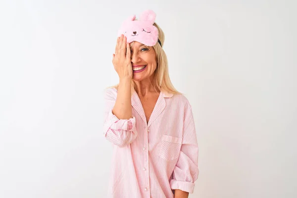 Middle age woman wearing sleep mask and pajama over isolated white background covering one eye with hand, confident smile on face and surprise emotion.