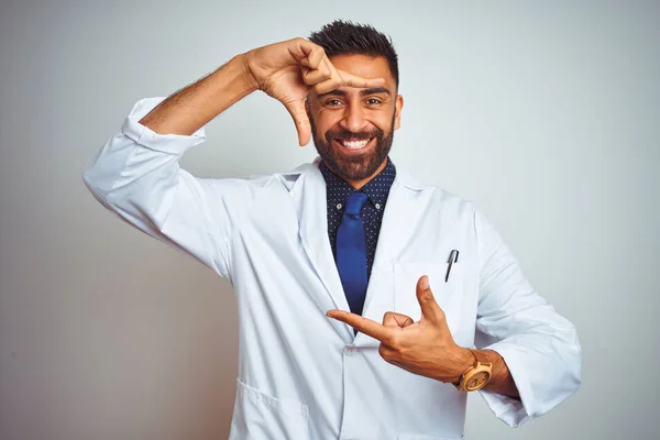 Young indian doctor man standing over isolated white background smiling making frame with hands and fingers with happy face. Creativity and photography concept.