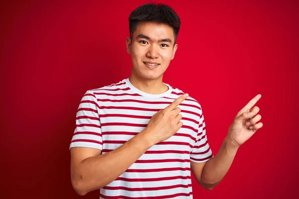 若いですアジアの中国人男性を身に着けていますストライプTシャツを身に着けています独立した赤い背景笑顔とカメラを見て2本の手と指で側面を指して — ストック写真