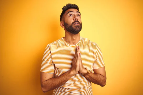 Young indian man wearing t-shirt standing over isolated yellow background begging and praying with hands together with hope expression on face very emotional and worried. Asking for forgiveness. Religion concept.