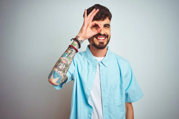 Young man with tattoo wearing blue shirt standing over isolated white background doing ok gesture with hand smiling, eye looking through fingers with happy face.