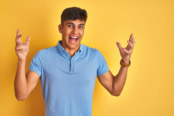 青いポロを身に着けている若いインド人男性は 孤立した黄色の背景の上に立って 腕を上げ 目を開けて興奮して成功に驚きました 勝者のコンセプト — ストック写真