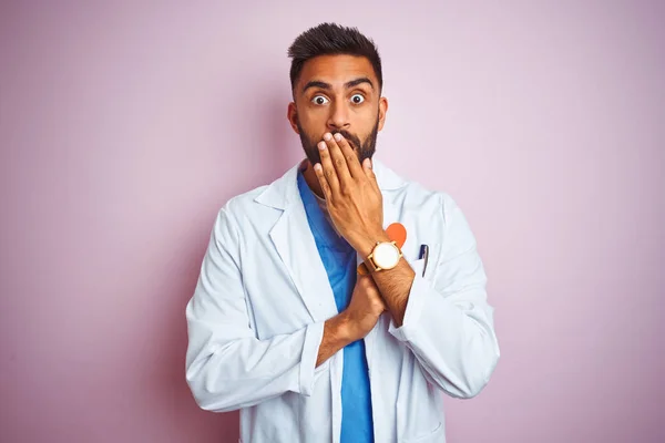 Young indian doctor man holding paper heart standing over isolated pink background cover mouth with hand shocked with shame for mistake, expression of fear, scared in silence, secret concept