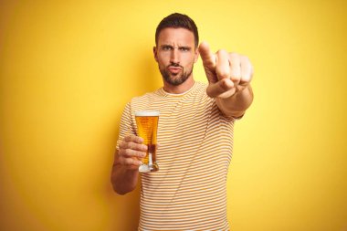 Genç yakışıklı adam, izole edilmiş sarı arka planda yarım litre bira içerek kamerayı işaret ediyor ve sen, el işareti, ön taraftan pozitif ve kendinden emin bir jest.