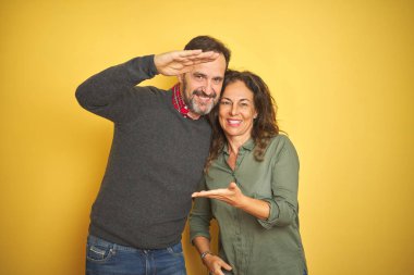 Güzel orta yaşlı çift, izole edilmiş sarı arka planda. Ellerinde büyük ve büyük boy işareti var. Ölçüm sembolü. Kameraya bakıp gülümsüyor. Ölçüm konsepti.