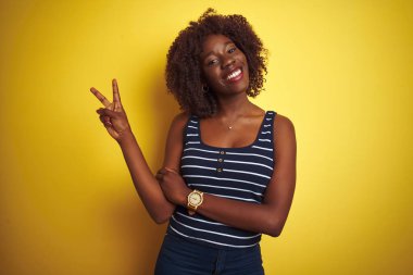 İzole edilmiş sarı arka plan üzerinde çizgili tişört giyen Afrikalı genç bir kadın kameraya gülümseyen ve zafer işareti yapan. İki numara..