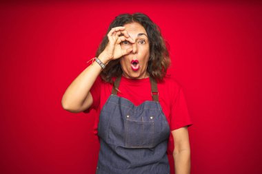 Kırmızı izole edilmiş arka planda önlük giyen orta yaşlı bir kadın şaşırmış bir yüz ve parmaklarından bakan bir gözle iyi bir jest yapıyor. İnançsız ifade.