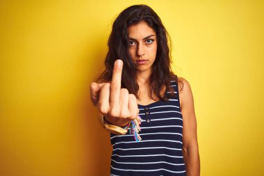 Sarı arka plan üzerinde duran çizgili tişört giyen genç ve güzel bir kadın orta parmak gösteriyor, kaba ve kaba bir ifade takınıyor.
