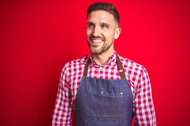 Kırmızı izole edilmiş arka planda dükkan sahibi üniforması giyen yakışıklı bir adam yüzünde gülümsemeyle, doğal bir ifadeyle yan yana bakıyor. Kendinden emin gülen.