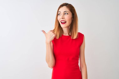 Kırmızılı iş kadını zarif kırmızı elbiseler giyiyor. İzole edilmiş beyaz arka planda duruyor. Gülümsüyor, gülümsüyor ve başparmağıyla yan tarafa bakıyor..