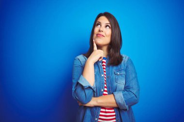 Genç, güzel, esmer kadın mavi kot ceket giyiyor ve mavi izole edilmiş arka plan üzerinde elleri çenesinde soru sormayı, düşünceli yüz ifadesini düşünüyor. Düşünceli bir yüzle gülümsüyor. Şüphe kavramı.