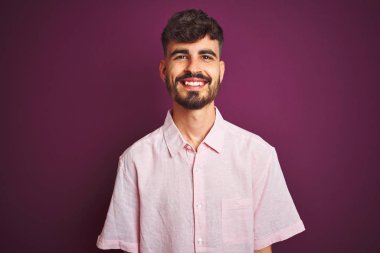 Yüzünde mutlu ve havalı bir gülümseme olan, izole edilmiş mor arka planda duran pembe tişörtlü, dövmeli genç adam. Şanslı kişi..