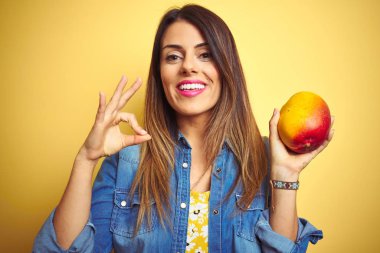 Genç ve güzel bir kadın sarı arka planda taze mango yiyor tamam işareti yapıyor parmaklarıyla, mükemmel bir sembol.