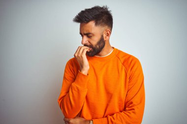 İzole edilmiş beyaz arka planda turuncu süveter giyen genç Hintli adam elleri ağız yiyen tırnaklarla stresli ve gergin görünüyor. Endişe sorunu.