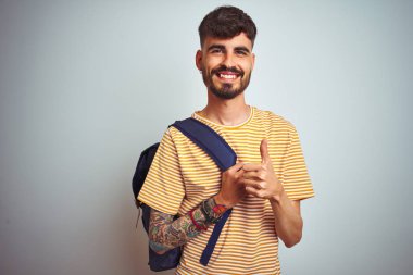 Sırt çantasında dövmesi olan genç bir öğrenci, izole edilmiş beyaz arka planda duruyor. Kocaman gülümsüyor ve gülümsüyor.