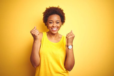 Beauitul Afro-Amerikan kadını izole edilmiş sarı arka plan üzerine yaz tişörtü giyerek kollarını kaldırarak ve açık gözlerle başarıyı kutluyor ve şaşırıyor. Kazanan konsept.