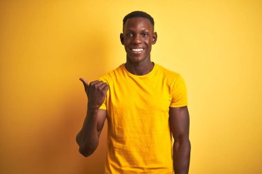 Genç, Afro-Amerikalı bir adam, günlük tişörtler giymiş, izole edilmiş sarı arka planda dikiliyor, gülümsüyor ve başparmağıyla yan tarafa bakıyor..