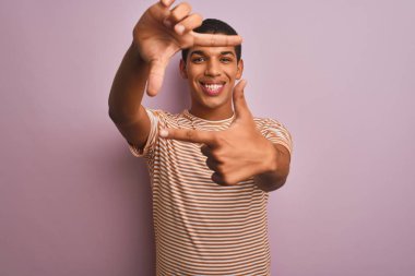 İzole edilmiş pembe arka planda duran çizgili tişört giyen yakışıklı Arap adam gülümsüyor ve elleri ve parmakları gülen yüzlü bir çerçeve yapıyor. Yaratıcılık ve fotoğrafçılık kavramı.