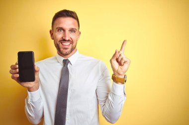 Genç yakışıklı iş adamı, sarı izole edilmiş arka plan üzerinde akıllı telefon ekranı gösteriyor.