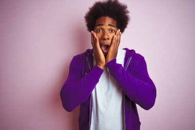Mor kazak giyen genç Afro-Amerikalı adam pembe arka planda duruyor. Korkmuş ve şok olmuş, şaşırmış ve şaşırmış bir halde.