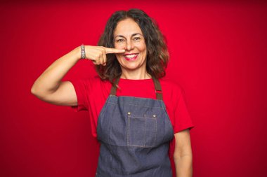 Kırmızı izole arka planda önlük giyen orta yaşlı bir kadın. El parmağı ve burunla işaret ediyor. Neşeli gülümsüyor. Güzellik konsepti