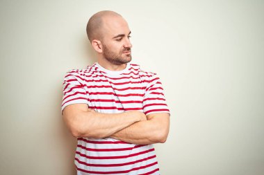 Sakallı genç ve kel bir adam beyaz izole edilmiş arka plan üzerine sıradan çizgili kırmızı tişört giyiyor kolları çapraz ve kendinden emin bir şekilde yana bakıyor.