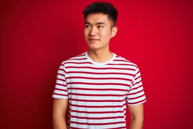 Asyalı genç bir Çinli, çizgili tişört giyiyor. İzole edilmiş kırmızı arka planda duruyor. Gülümsüyor ve başka tarafa bakıyor..
