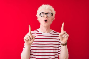 Genç albino sarışın adam çizgili tişört ve gözlüklerle izole edilmiş kırmızı arka planda şaşkın şaşkın yukarı bakıyor ve parmaklarıyla işaret ediyor ve kollarını kaldırıyor..