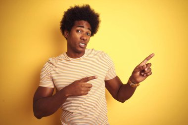 Afro saçlı, çizgili tişörtlü, izole edilmiş sarı arka planda dikilen Amerikalı bir adam. Endişeli ve gergin bir tarafı var. Endişeli ve şaşırmış bir ifadesi var.