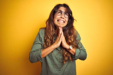 Yeşil gömlek ve gözlük takan genç güzel bir kadın izole edilmiş sarı arka planda yalvarıyor ve dua ediyor. Yüzünde umut ifadesi olan çok duygusal ve endişeli bir ifade var. Af diliyor. Din kavramı.