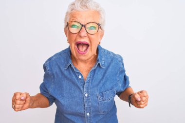 Yaşlı, gri saçlı, kot gömlek giyen ve gözlüklü bir kadın izole edilmiş beyaz arka plan üzerine kızgın ve öfkeli çığlık atan, öfkeyle bağıran bir kadın. Öfke ve agresif kavram.