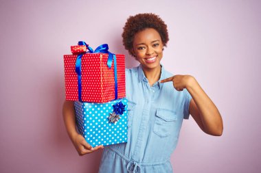 Afro-Amerikalı bir kadın pembe izole edilmiş arka planda doğum günü hediyelerini tutuyor. Sürpriz yüz ifadesiyle kendini gösteriyor.