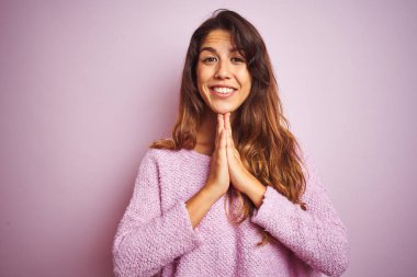 Pembe arka plan üzerinde duran genç ve güzel bir kadın ellerini birleştirip af diliyor kendinden emin bir şekilde gülümsüyor..