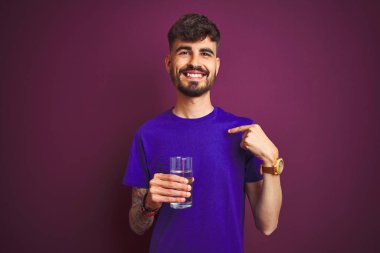 Dövmeli genç adam izole edilmiş mor arka planda duran su bardağı içerken kendini işaret eden sürpriz yüzlü adam.