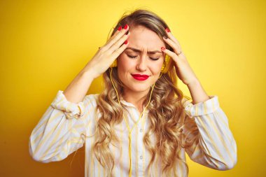 Genç ve güzel bir kadın, sarı arka plan üzerinde kulaklık kullanarak müzik dinliyor. Ağrı ve migrenden dolayı çaresiz ve stresli bir baş ağrısı çekiyor. Ellerini başının üstüne koy..