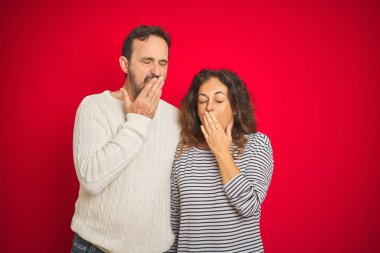 Güzel orta yaşlı çift izole edilmiş kırmızı arka plan üzerine kışlık süveter giyiyorlar. Ağızlarını eliyle kapatmaktan sıkıldılar. Huzursuz ve uykulu..