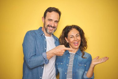 Güzel orta yaşlı çift, izole edilmiş sarı arka planda kot gömlek giyiyorlar. Kameraya hayret ediyorlar ve gülümsüyorlar. Elleriyle ve parmaklarıyla gösteriyorlar..